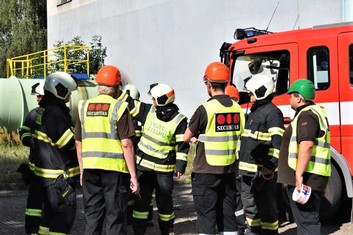Velitel zásahu ukazuje ostatním podnikovým hasičům a členům ostrahy, jak daleko od objektu s  balíčkem mají natáhnout zákazovou pásku.